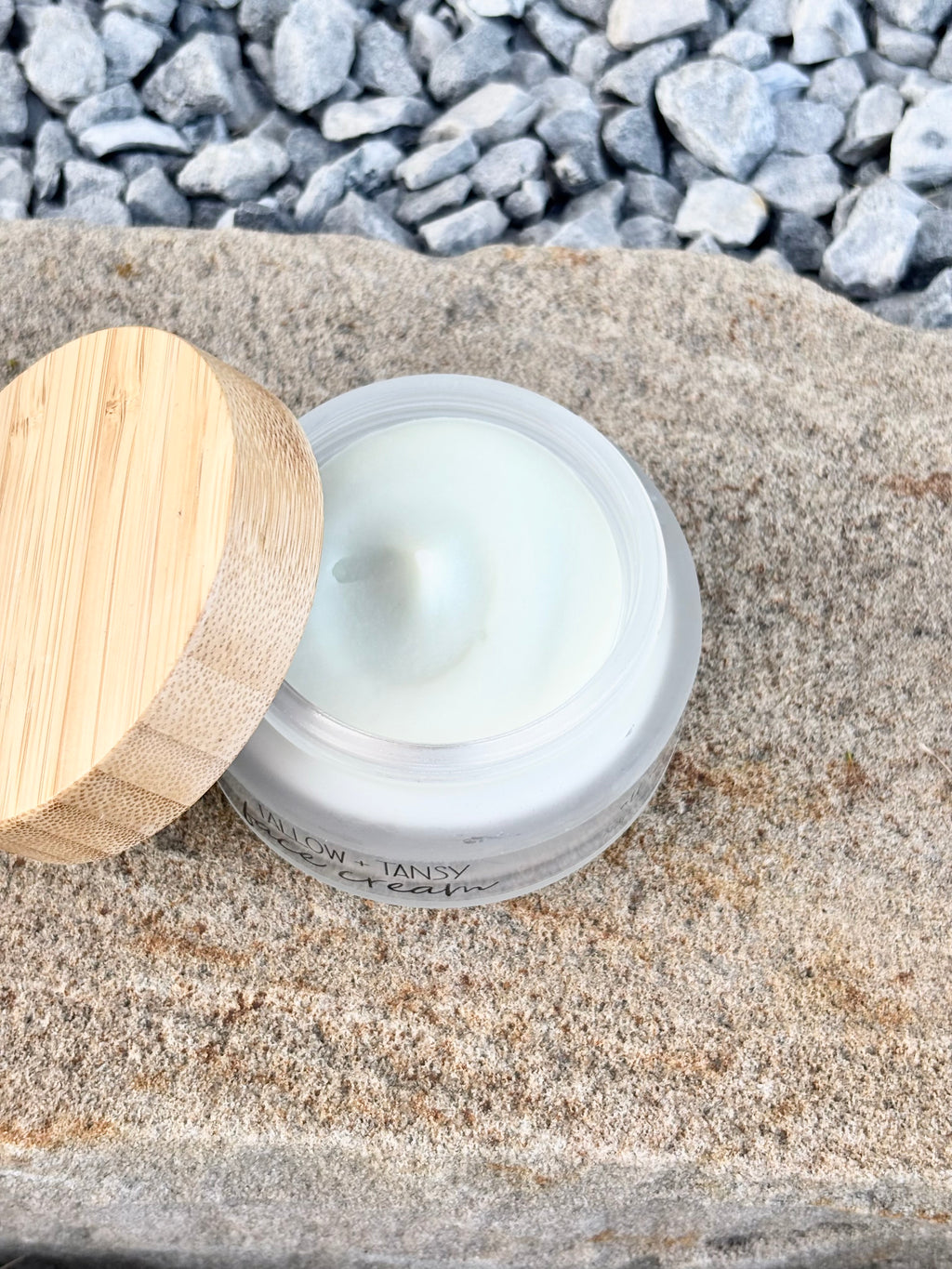 An open jar of pale greenish-white cream or balm with a light wood lid resting beside it on a stone surface, with small gray pebbles in the background.