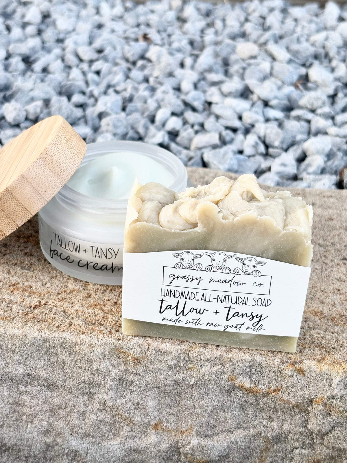 A handmade soap bar labeled 'tallow + tansy' by Grassy Meadow Co, shown next to its open jar with a bamboo lid on a stone surface with gravel in the background.