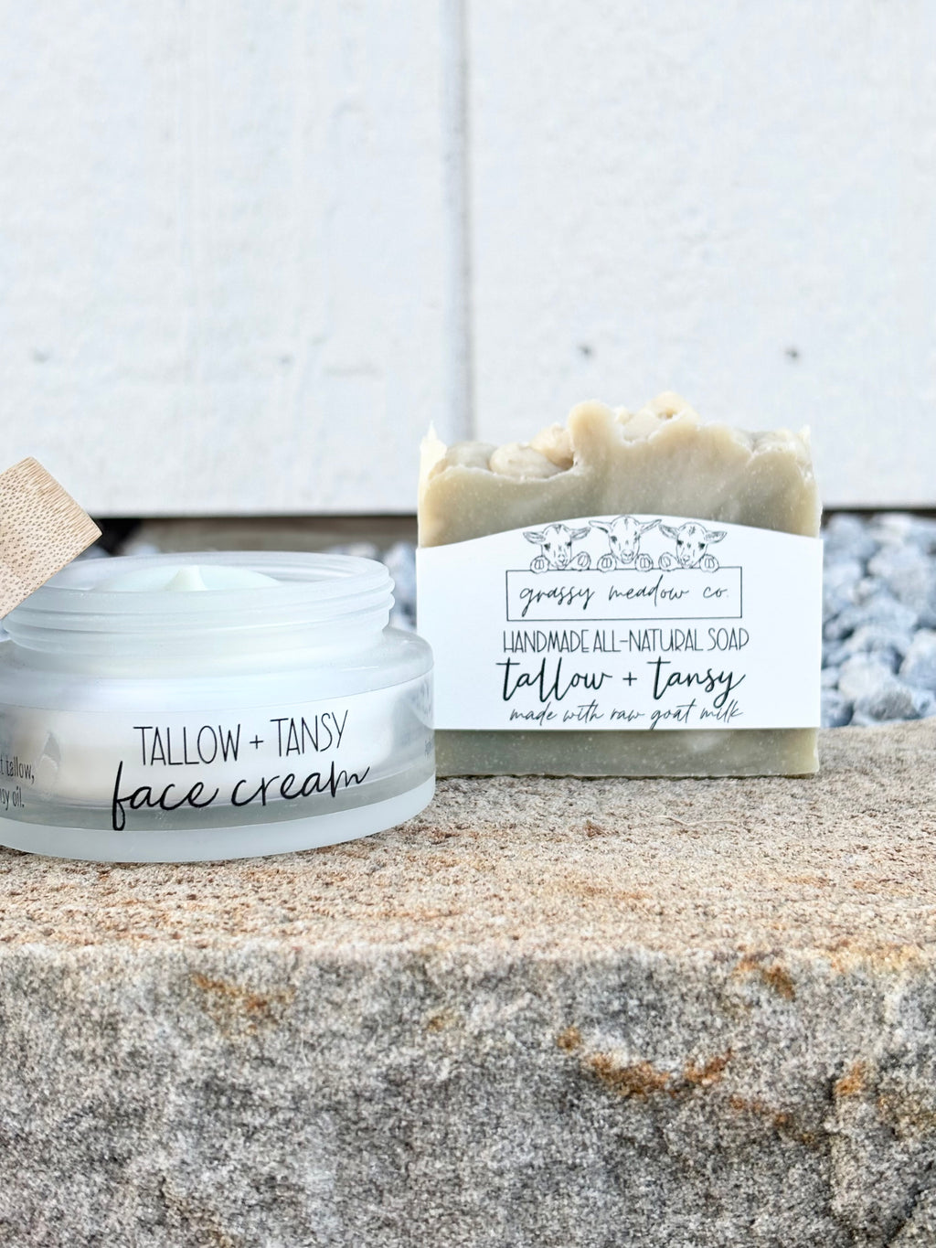 A jar of Tallow + Tansy face cream and a handmade soap bar from Grassy Meadow Co, styled on a stone surface against a white wooden background.