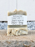 A handmade soap bar wrapped in a white paper label reading 'grassy meadow co' and 'tallow + tansy made with raw goat milk', standing upright on a stone surface with a blurred outdoor background.