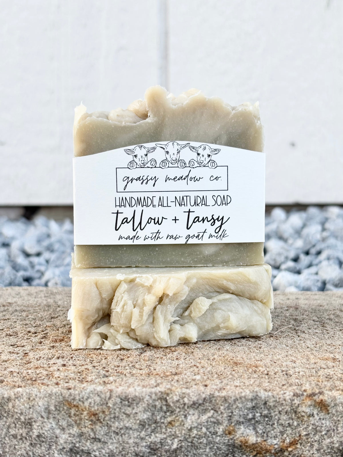 A handmade soap bar wrapped in a white paper label reading 'grassy meadow co' and 'tallow + tansy made with raw goat milk', standing upright on a stone surface with a blurred outdoor background.