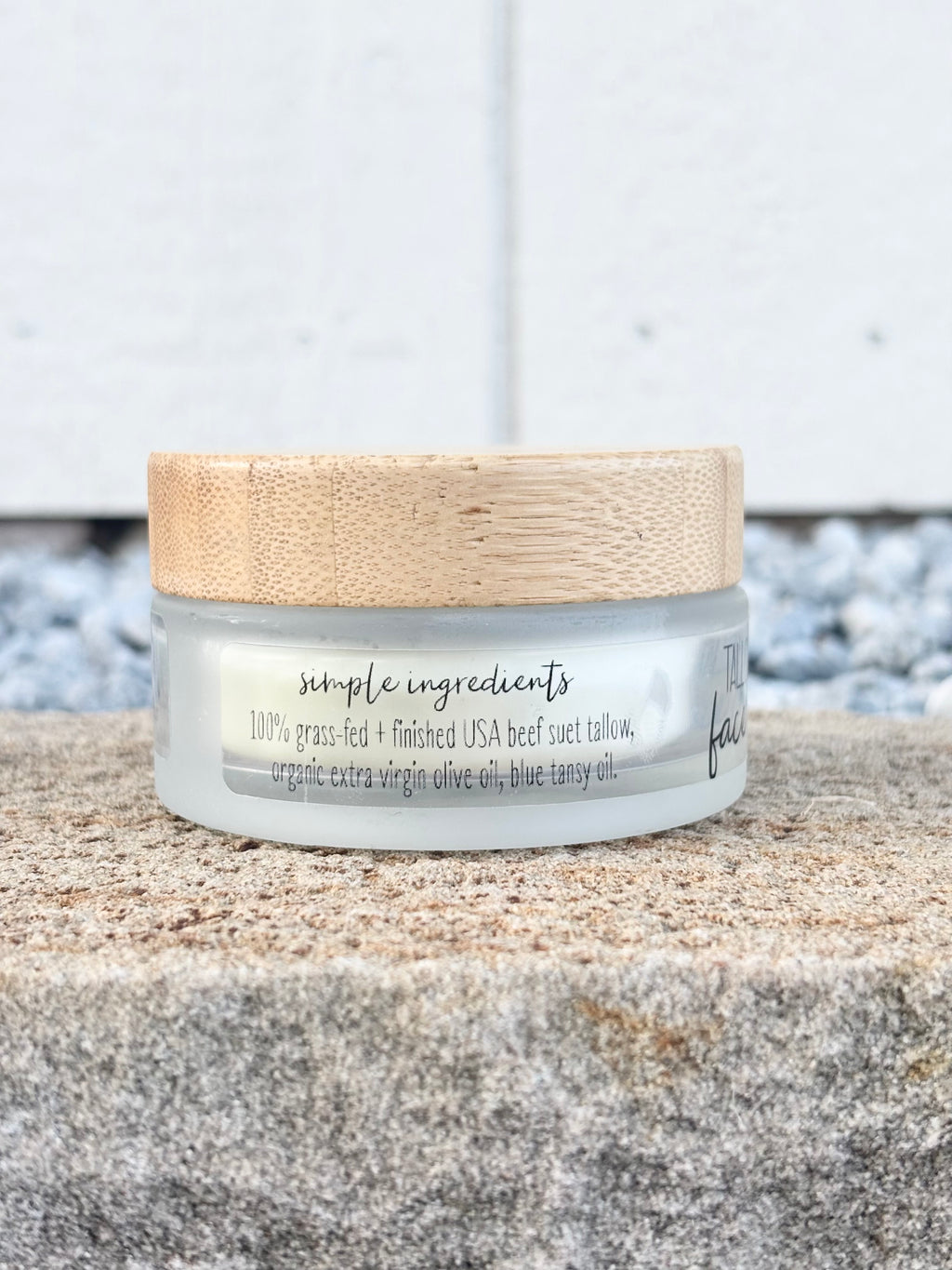 A small clear cosmetic jar with a light wood lid sits on a stone surface against a white wall background. The label reads 'simple ingredients' and '100% grass-fed + finished USA beef suet tallow, organic extra virgin olive oil, blue tansy oil.