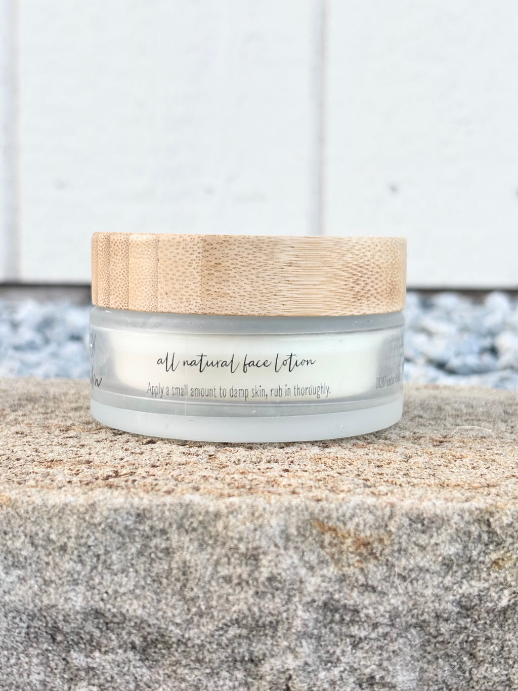A small jar of face lotion with a bamboo-style lid sits on a stone surface in front of a white paneled wall.