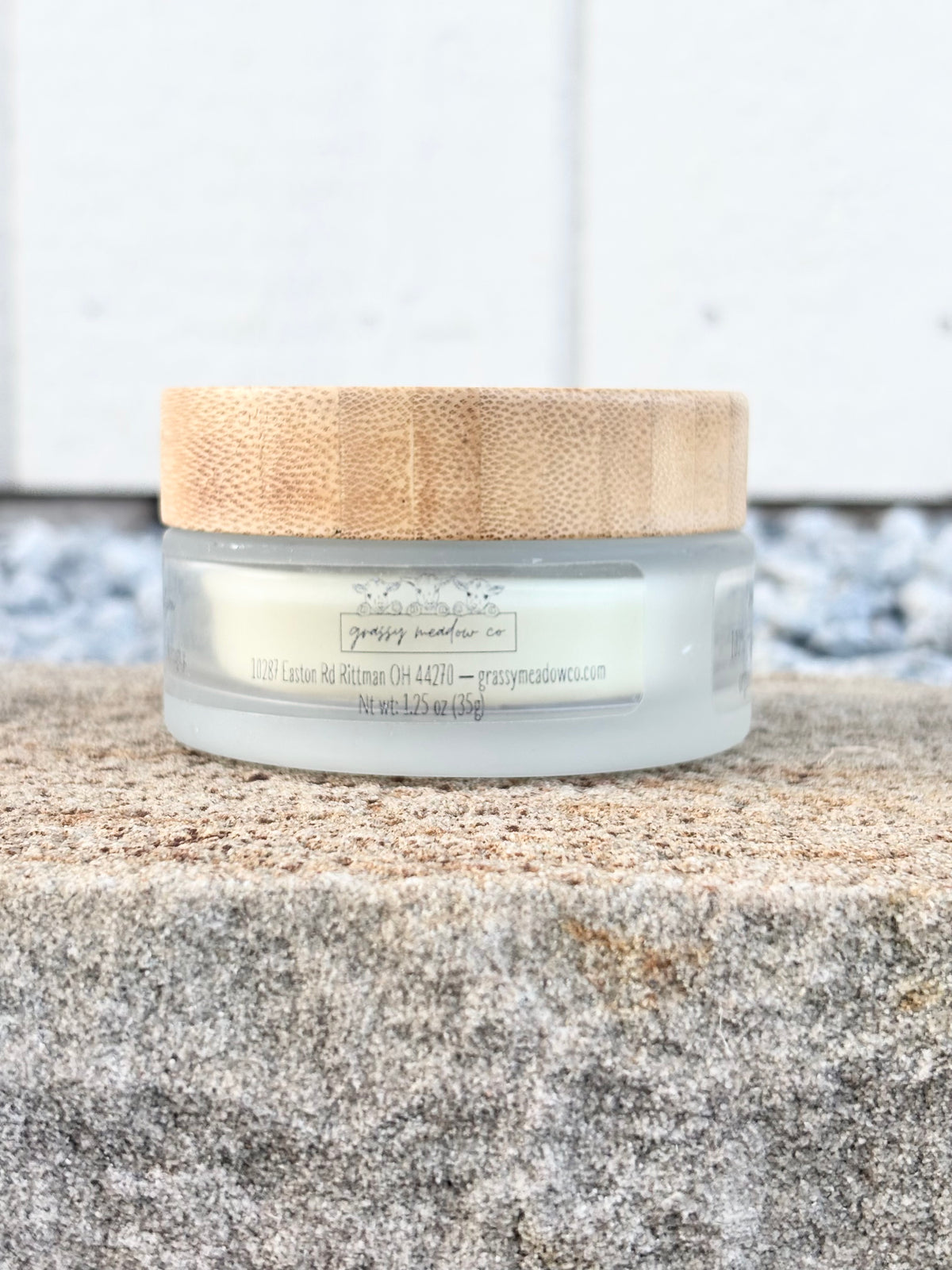 A small frosted glass jar of body butter with a light natural wood lid, resting on a textured stone surface against a white paneled background.