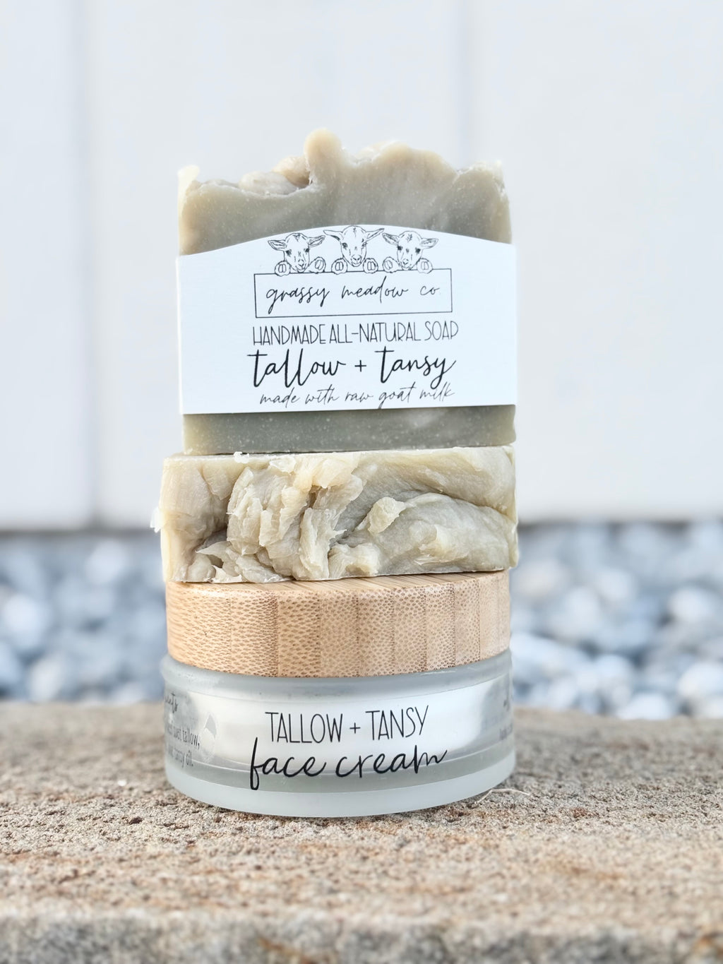 A stacked set of skincare products from Grassy Meadow Co, including a handmade soap bar labeled 'Tallow + Tansy' and a jar of 'Tallow + Tansy face cream' with a bamboo lid, set on a sandy surface with blurred pebbles and white siding in the background.